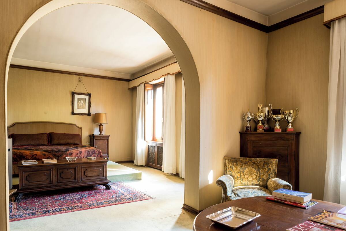 A bedroom with a bed, a nightstand, a lamp, a chair, a table, and trophies on a cabinet.