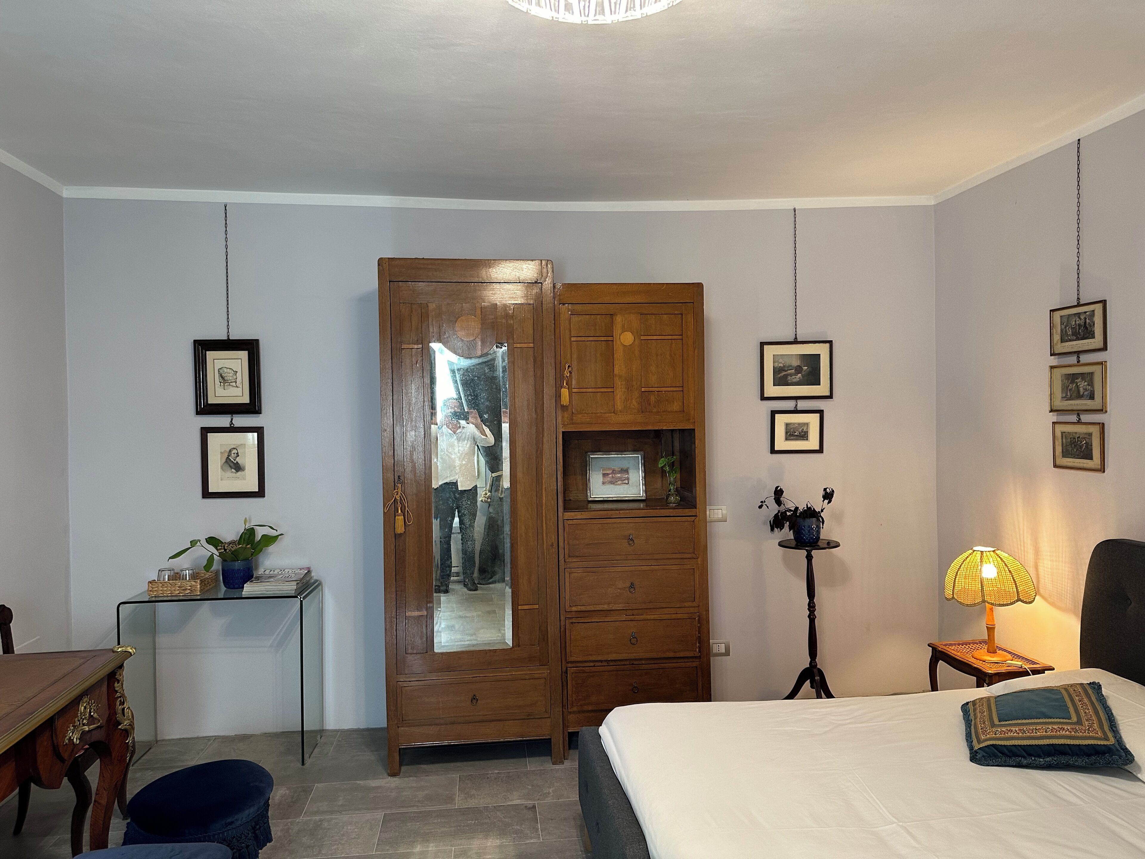 A bedroom with a bed, a wooden wardrobe, a desk with a plant, and framed pictures on the wall.