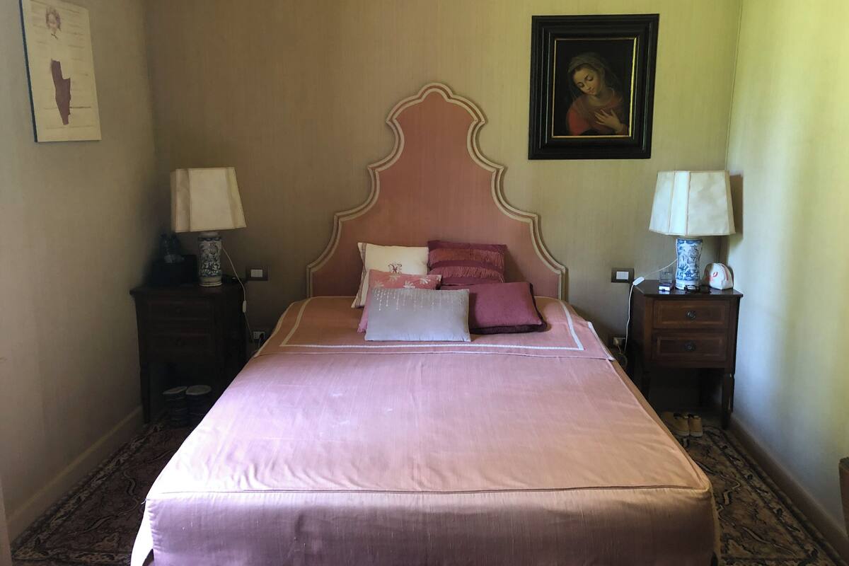 A bedroom with a large bed, two bedside tables, a nightstand, a framed picture on the wall, and a carpeted floor.