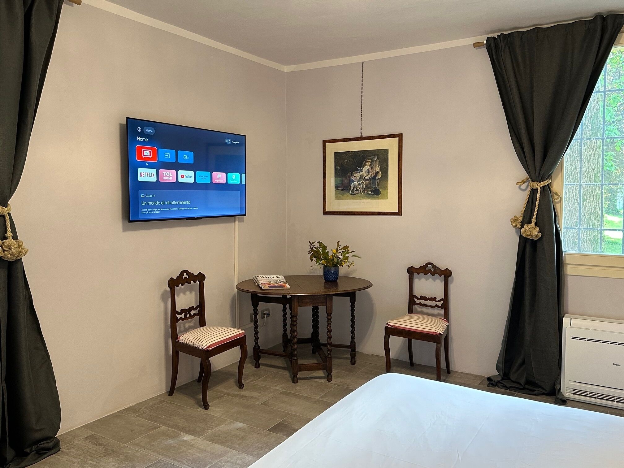 Ett hotellrum med en säng, två trästolar, ett litet bord med en växt, en tv som är monterad på väggen och visar olika appar, och en inramad tavla ovanför bordet.