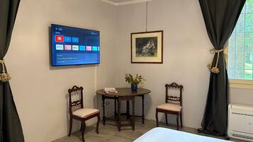 Ett hotellrum med en säng, två trästolar, ett litet bord med en växt, en tv som är monterad på väggen och visar olika appar, och en inramad tavla ovanför bordet.