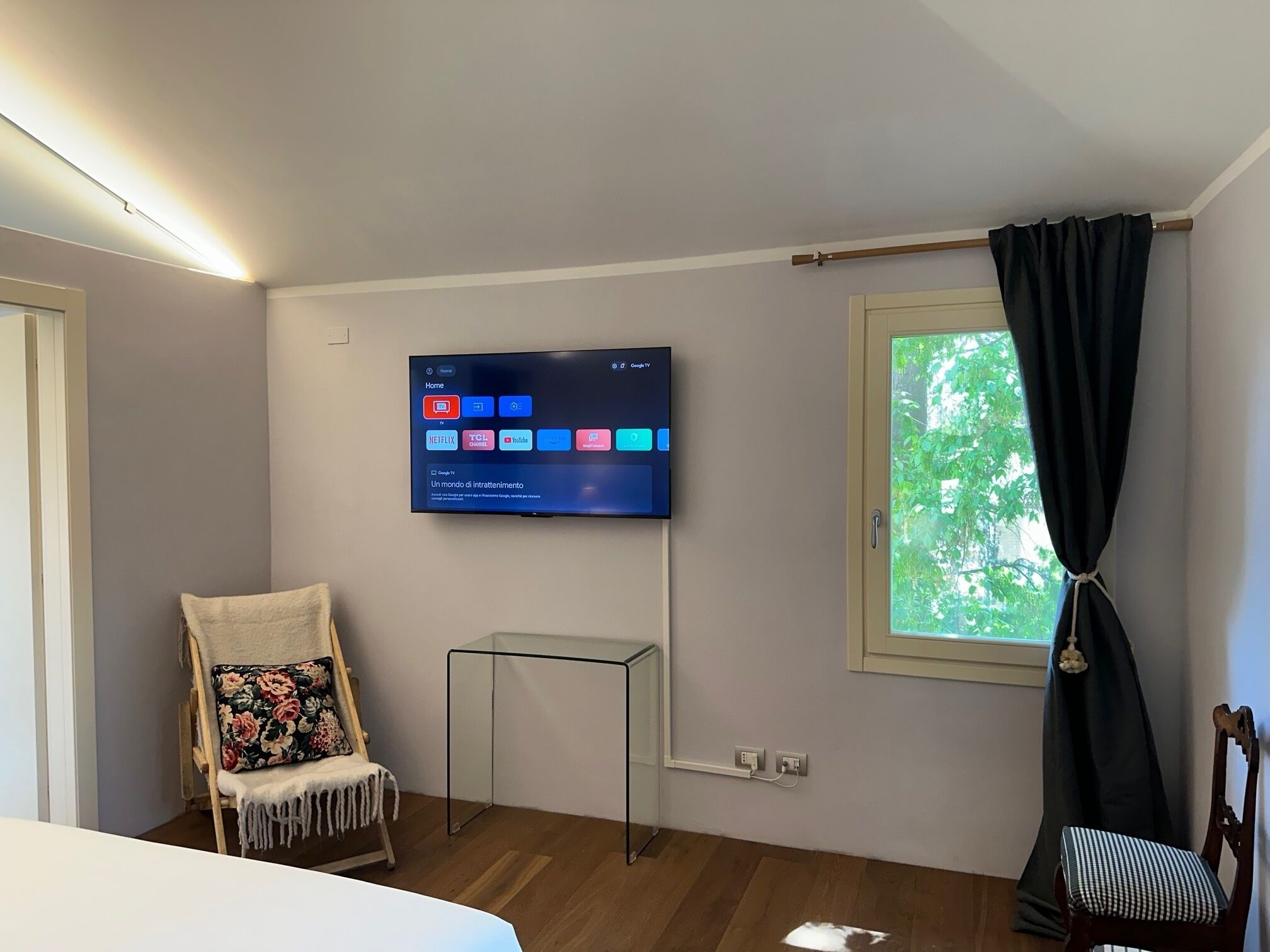 Hotelkamer met een flatscreen televisie aan de muur waarop verschillende apps worden getoond, een klein tafeltje, een stoel met een bloemenkussen, een raam met gordijn en een gedeeltelijk zichtbaar bed.