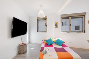Una camera da letto moderna con una grande TV a schermo piatto, un letto con lenzuola colorate, un piccolo comodino e una finestra con persiane.
