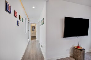 Un corridoio con una TV a schermo piatto montata sulla parete, un tavolino di legno con il telecomando e opere d'arte incorniciate al muro.