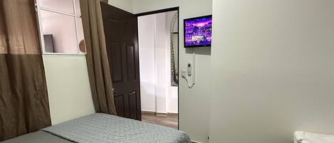 Un dormitorio con una cama, un televisor montado en la pared y una puerta que da a otra habitación.