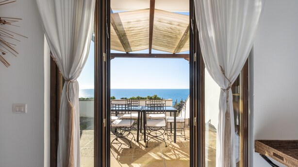 Vista a través de un ventanal a un patio exterior con una mesa y sillas, una sombrilla blanca y una vista despejada del mar.