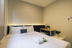 A hotel room with a neatly made bed, a bedside table with a lamp, a small plant, and a wall-mounted light switch. - Deco Lux Hotel (Bangkok)