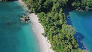 An aerial view of a beach with clear turquoise waters, surrounded by dense green foliage and palm trees. - Villa Zest 5 bedroom Villa • Rooftop Infinity Pool & Ocean Views (Quepos)