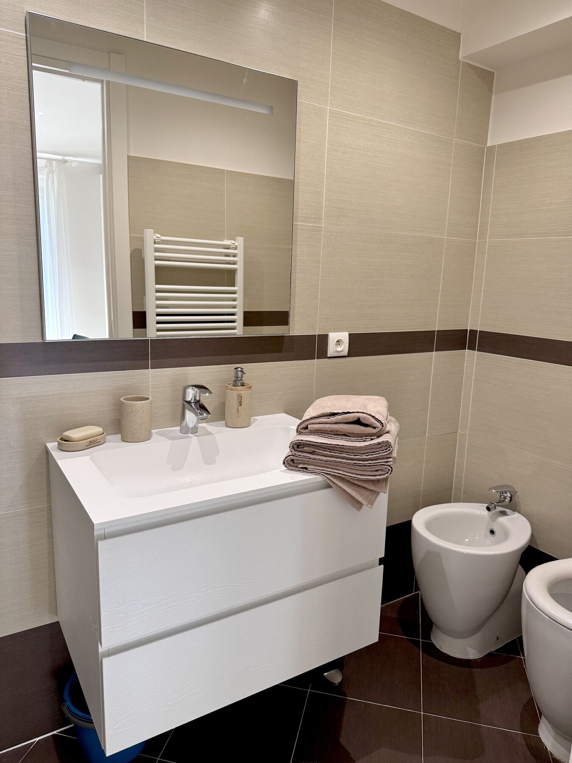 A modern bathroom with a white vanity, a large mirror, a bidet, and a towel rack.
