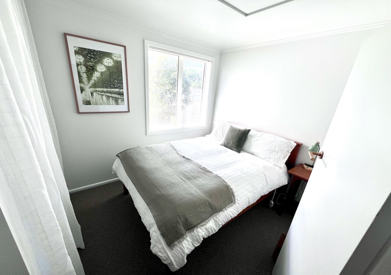 Une chambre à coucher bien rangée, avec un lit, une table de chevet, une lampe et une fenêtre équipée de stores.