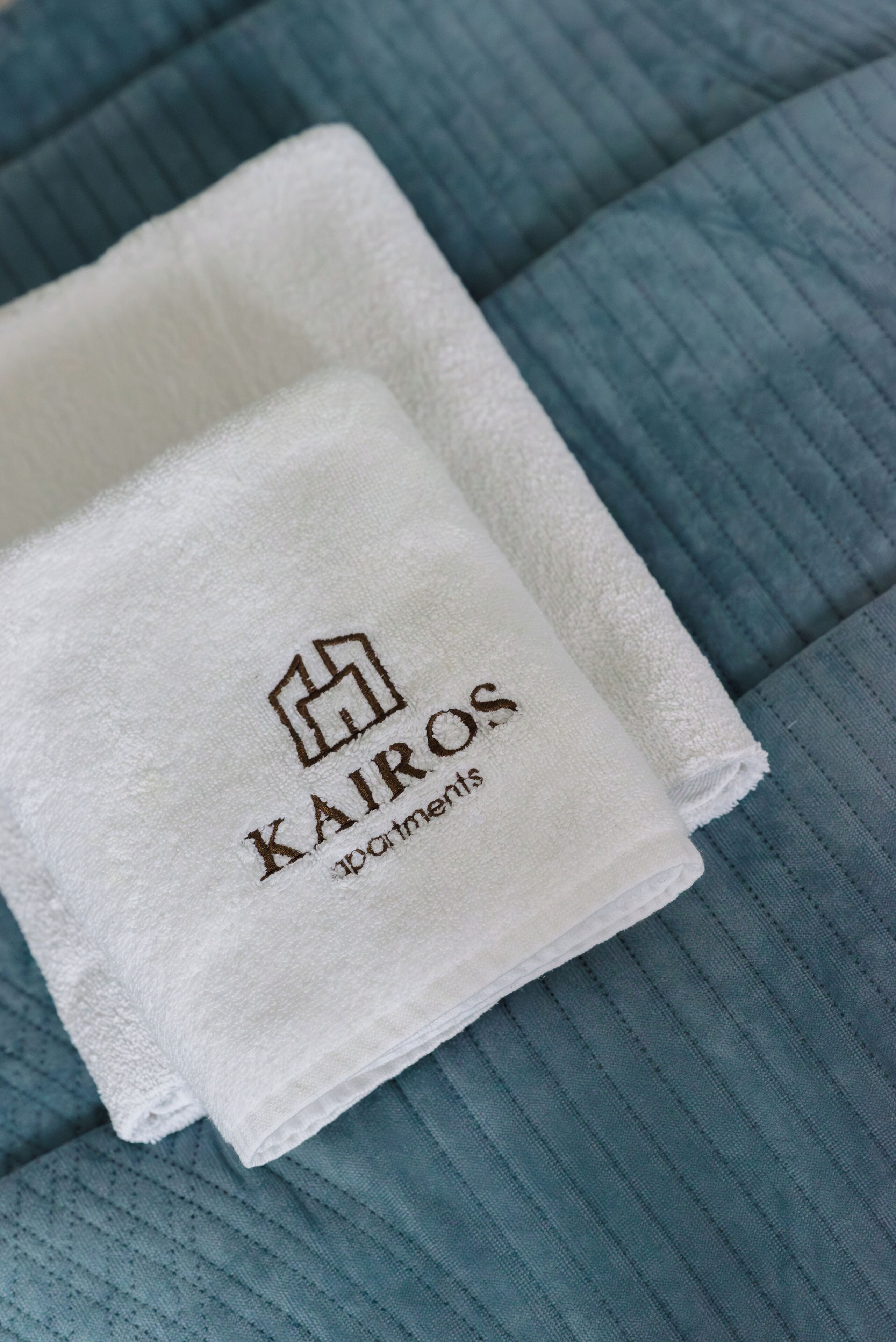 Una toalla blanca doblada con el logotipo y el nombre 'Kairos Apartments', sobre una superficie de tela azul con textura.