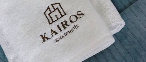 Una toalla blanca doblada con el logotipo y el nombre 'Kairos Apartments', sobre una superficie de tela azul con textura.