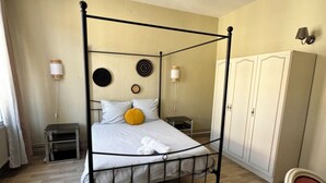 A four-poster bed with a white mattress and a single yellow pillow, a small bedside table, a wardrobe, and a chair in a room with wooden flooring and cream-colored walls. - 5 bedroom house and courtyard - 1930's Roubaix (Roubaix)