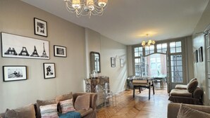 A spacious living room with a herringbone wooden floor, a wooden coffee table, and a sofa with a blue throw. There are framed pictures on the walls, a chandelier, and a dining area in the background. - 5 bedroom house and courtyard - 1930's Roubaix (Roubaix)