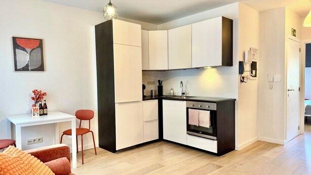 Een compacte keuken met witte kasten, een zwart aanrecht en een inbouwoven. Er is een klein eethoekje met een witte tafel en twee stoelen.