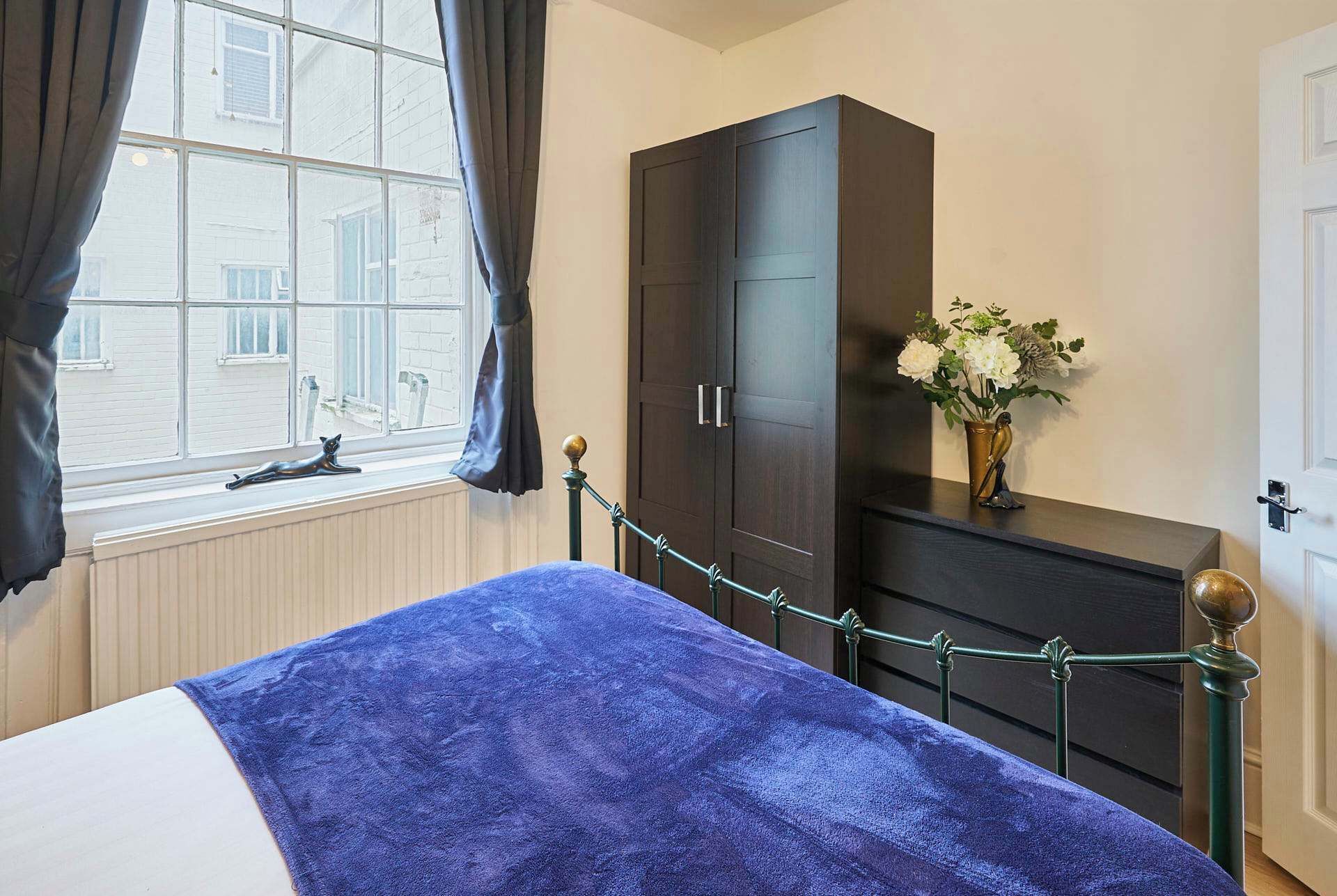 A bedroom with a bed, a wardrobe, a window with curtains, and a vase with flowers on a dresser.
