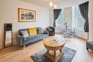 A modern living room with a sofa, a coffee table, and a dining area with chairs and a table. - Host Stay Grand Parade Apartment (Brighton)