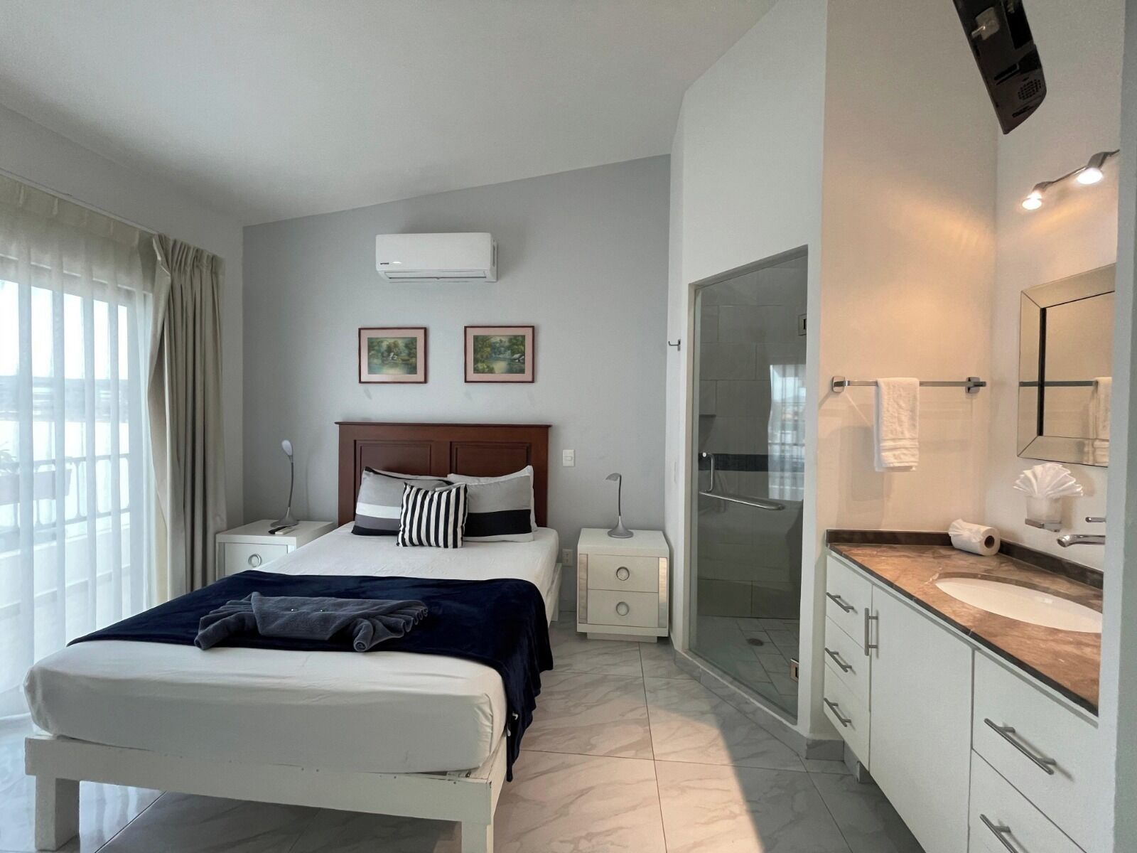 A modern hotel room with a large bed, bedside tables, a shower, a vanity with a mirror, and two framed pictures on the wall.