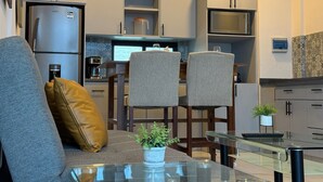 A modern kitchen with a refrigerator, microwave, and coffee maker. There is a dining area with a glass table and two chairs. A sofa with a brown pillow is in the foreground. - The Residences Apart-Hotel By Morgan (Cabo San Lucas)
