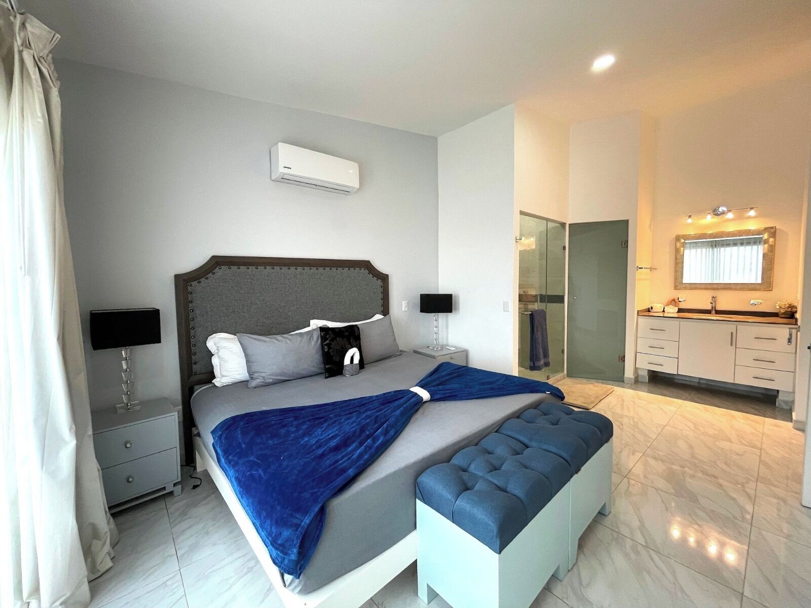 A modern bedroom with a large bed, bedside tables, a bench, a bathroom with a mirror and sink, and an air conditioning unit.