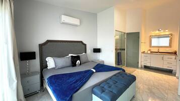 A modern bedroom with a large bed, bedside tables, a bench, a bathroom with a mirror and sink, and an air conditioning unit.