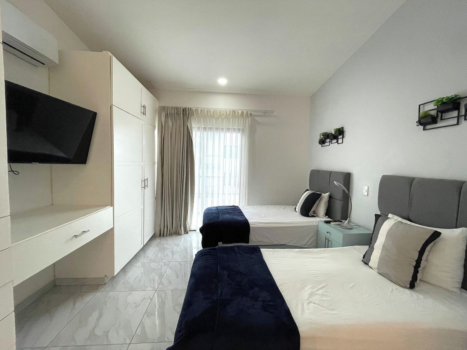 A modern hotel room with a bed, a TV, a desk, and a window with curtains.