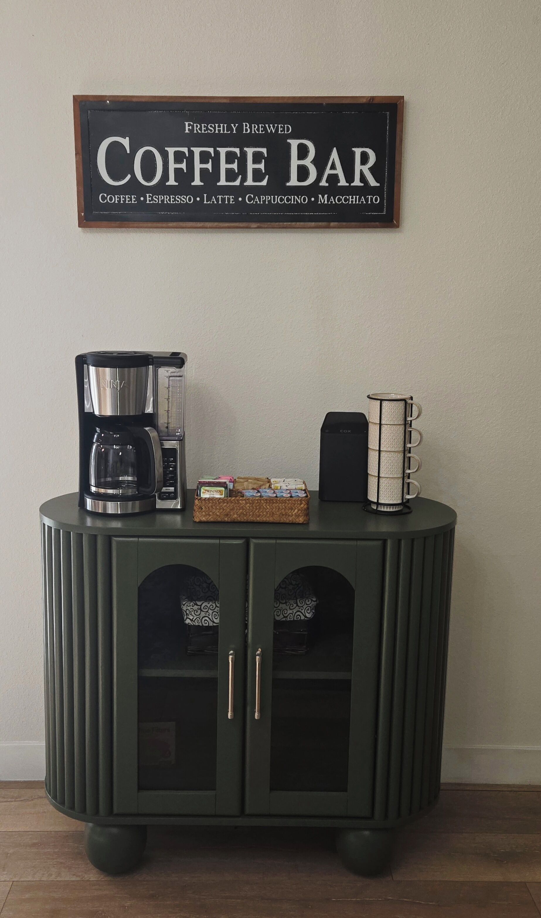 A dark green cabinet with a coffee maker, a box of coffee pods, and a coffee grinder on top.