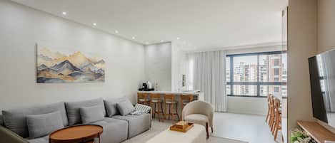 Una sala moderna con un sofá gris, una mesa de centro y un televisor. Hay un comedor con una mesa y sillas, y un ventanal con vistas a la ciudad.