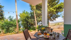 Zona pranzo all'aperto con tavolo apparecchiato per la colazione, immersa nel verde e con vista panoramica sul paesaggio.