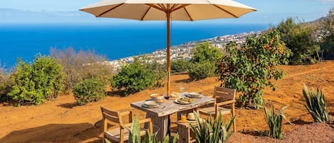 An outdoor dining setup with a table, chairs, and an umbrella overlooking a coastal landscape.