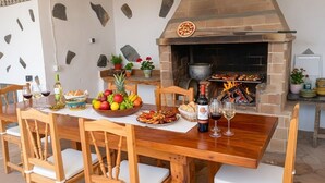 Un'area pranzo all'aperto con un forno a mattoni, sedie di legno, un tavolo apparecchiato con cibo e bevande e uno sfondo di parete in pietra.