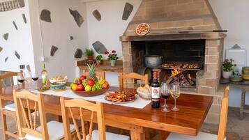 An outdoor dining area with a brick oven, wooden chairs, a table set with food and drinks, and a stone wall backdrop.