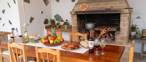 An outdoor dining area with a brick oven, wooden chairs, a table set with food and drinks, and a stone wall backdrop.