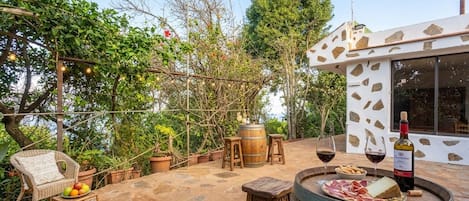 Une terrasse extérieure avec un fauteuil en osier, des tabourets en bois, un tonneau à vin et une bouteille de vin.