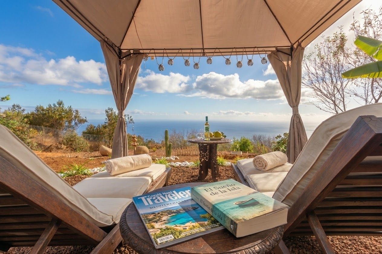 A covered outdoor seating area with lounge chairs and a table, overlooking a scenic landscape with a view of the sea and distant hills.