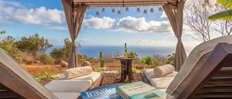 Un espace extérieur aménagé, abrité et doté de fauteuils et d’une table, offrant une vue panoramique sur un paysage magnifique, avec la mer et des collines lointaines en toile de fond.