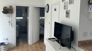 Et værelse med et fjernsyn på et stativ, en dør der fører til et andet rum, en lille hylde med genstande og en væg med billeder i rammer.