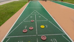 A shuffleboard table with a green playing surface and numbered scoring areas, set up under a wooden pergola with a stone building in the background. - DEC 27-Jan 3   89 per ngt.  COZY KING STUDIO! Enjoy Christmas Shows  in Branson (Branson)