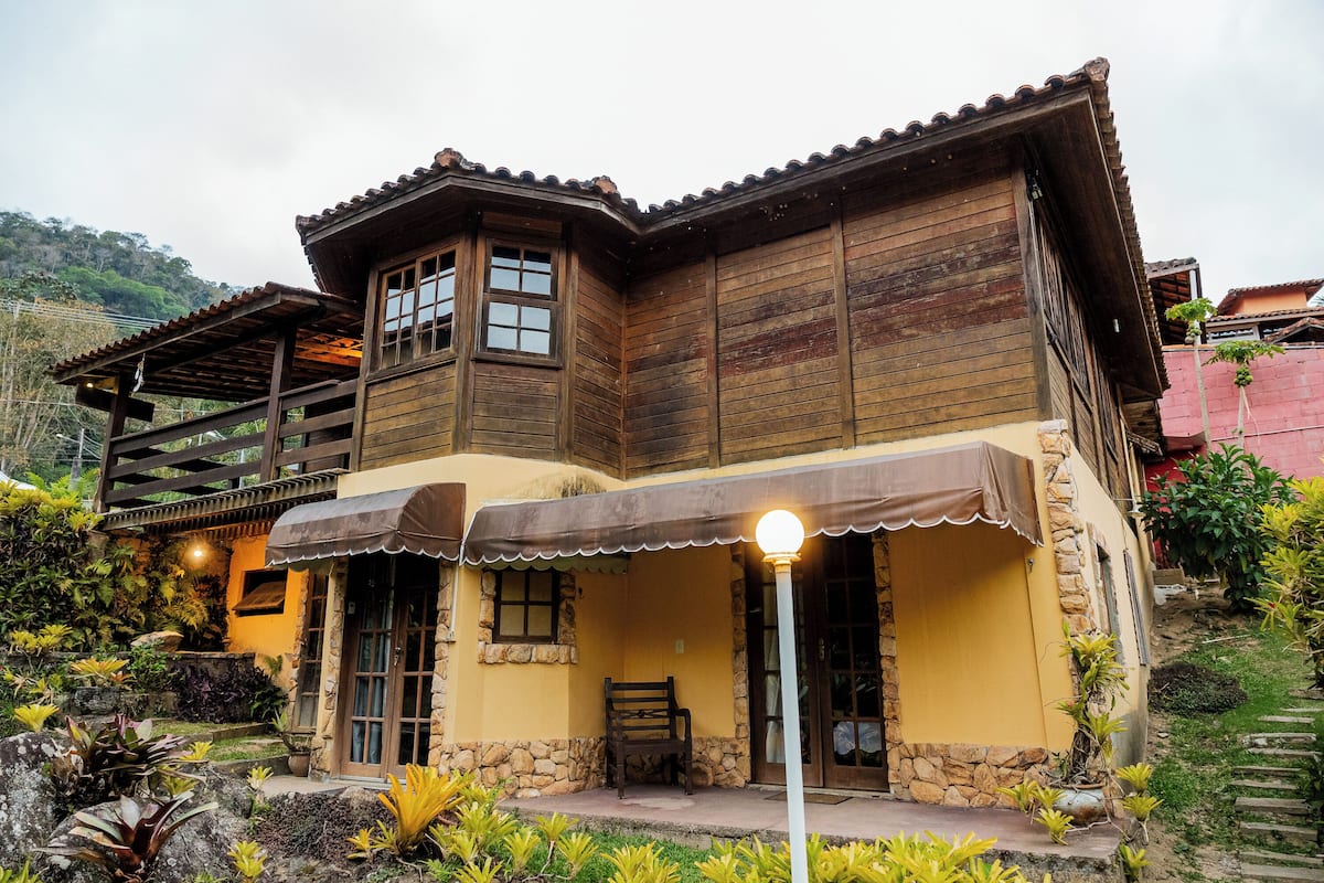 A two-story wooden house with a balcony, stone foundation, and a covered porch area.