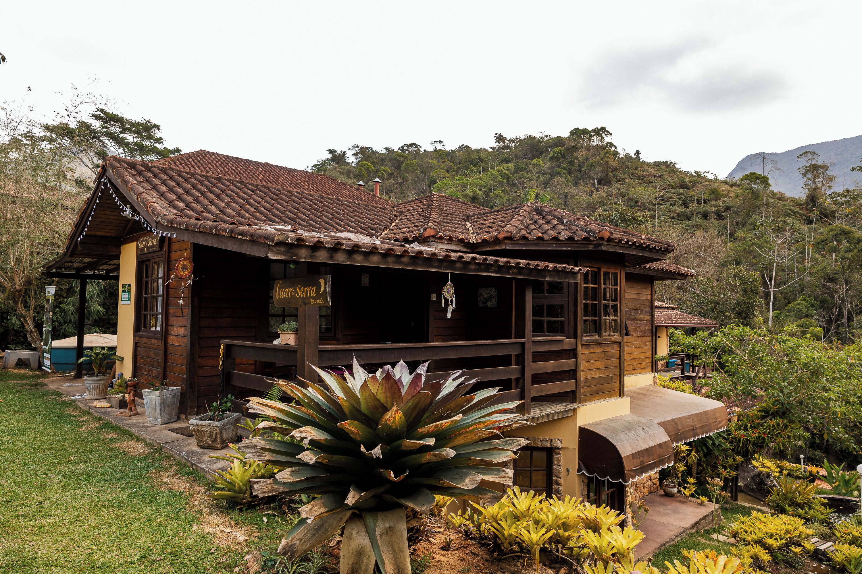 A rustic wooden building with a tiled roof, surrounded by lush greenery and a mountainous backdrop.