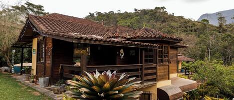 A rustic wooden building with a tiled roof, surrounded by lush greenery and a mountainous backdrop.