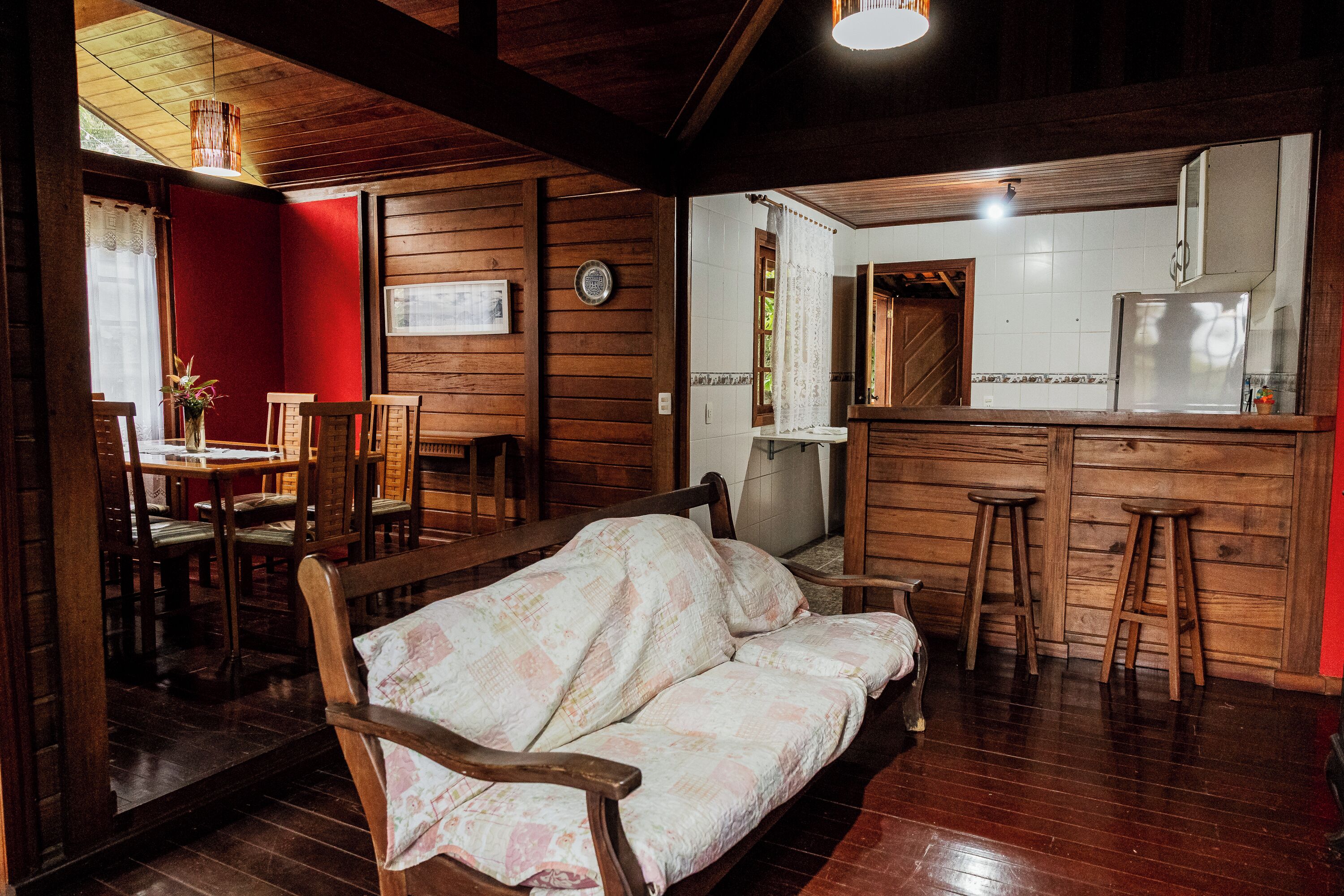 A cozy room with a wooden sofa, a dining table with chairs, a kitchen area with a refrigerator, and a red accent wall.