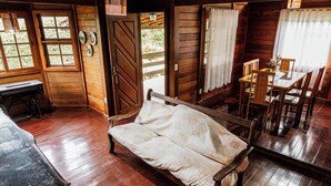 Interior de una cabaña de madera con una cama, una mesa de comedor con sillas y una ventana con cortinas.