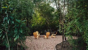 Un área de estar al aire libre con sillas de madera y una mesa, rodeada de abundante follaje y un camino de grava.