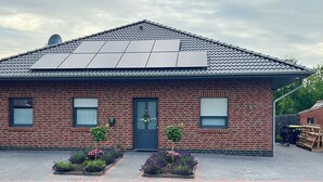 A brick house with a tiled roof, solar panels, a front door, two windows, and a small garden with flowers.
