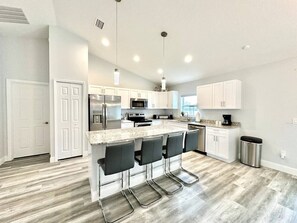 Una cocina moderna con una isla central, taburetes de bar, electrodomésticos de acero inoxidable y muebles de color blanco.