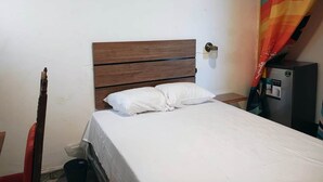 Een eenvoudige kamer met een houten bed, wit beddengoed, een klein nachtkastje, een wandlamp en een kleurrijk gordijn.