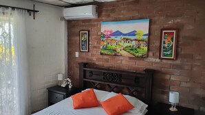 A hotel room with a brick wall, a bed with orange pillows, a painting of a village, and a bedside table with a lamp. - Lombardia Planes de Renderos - Casa de Piedra (San Salvador)