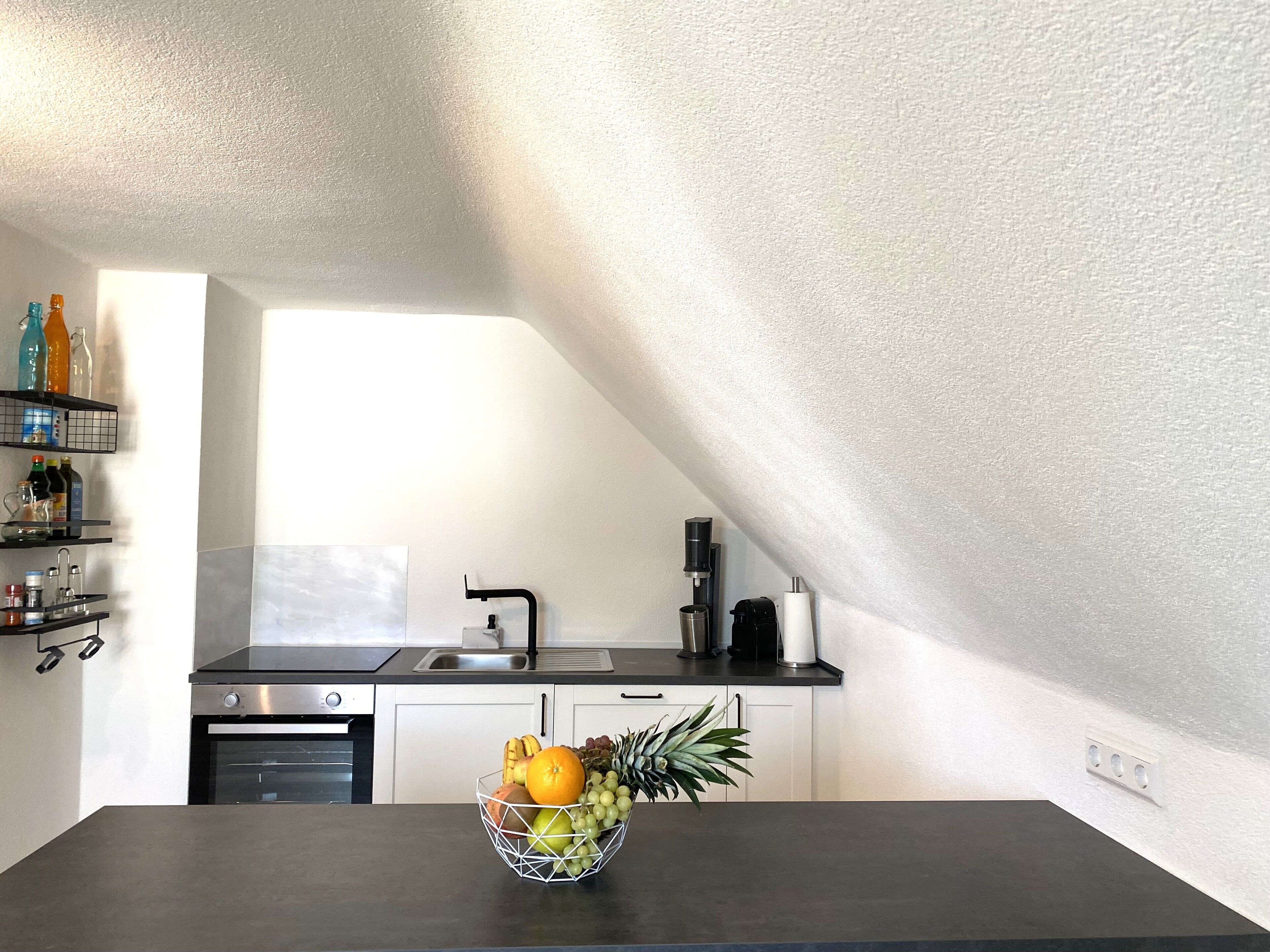 Eine Küche mit schwarzem Arbeitsplatz, einer Obstschale, einer Kaffeemaschine und einem Fenster.