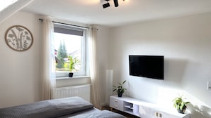 A modern bedroom with a sofa, a TV mounted on the wall, a window with a view of greenery outside, and a radiator under the window. - Modern feel-good apartment in the Illertal (Dettingen an der Iller)